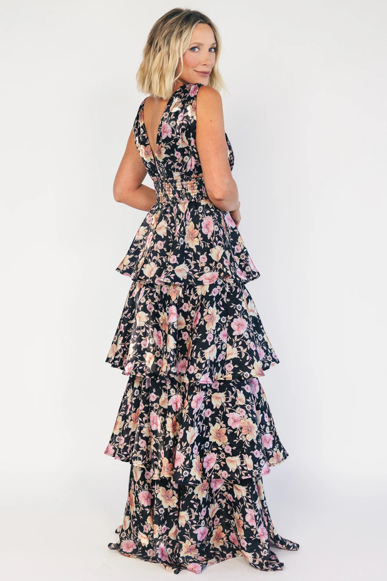 Model wearing a black floral maxi dress with a deep V-neckline, banded waist, and cascading tiered skirt. The dress features a black background with pink, yellow, and orange floral print, creating a flowing, romantic silhouette.