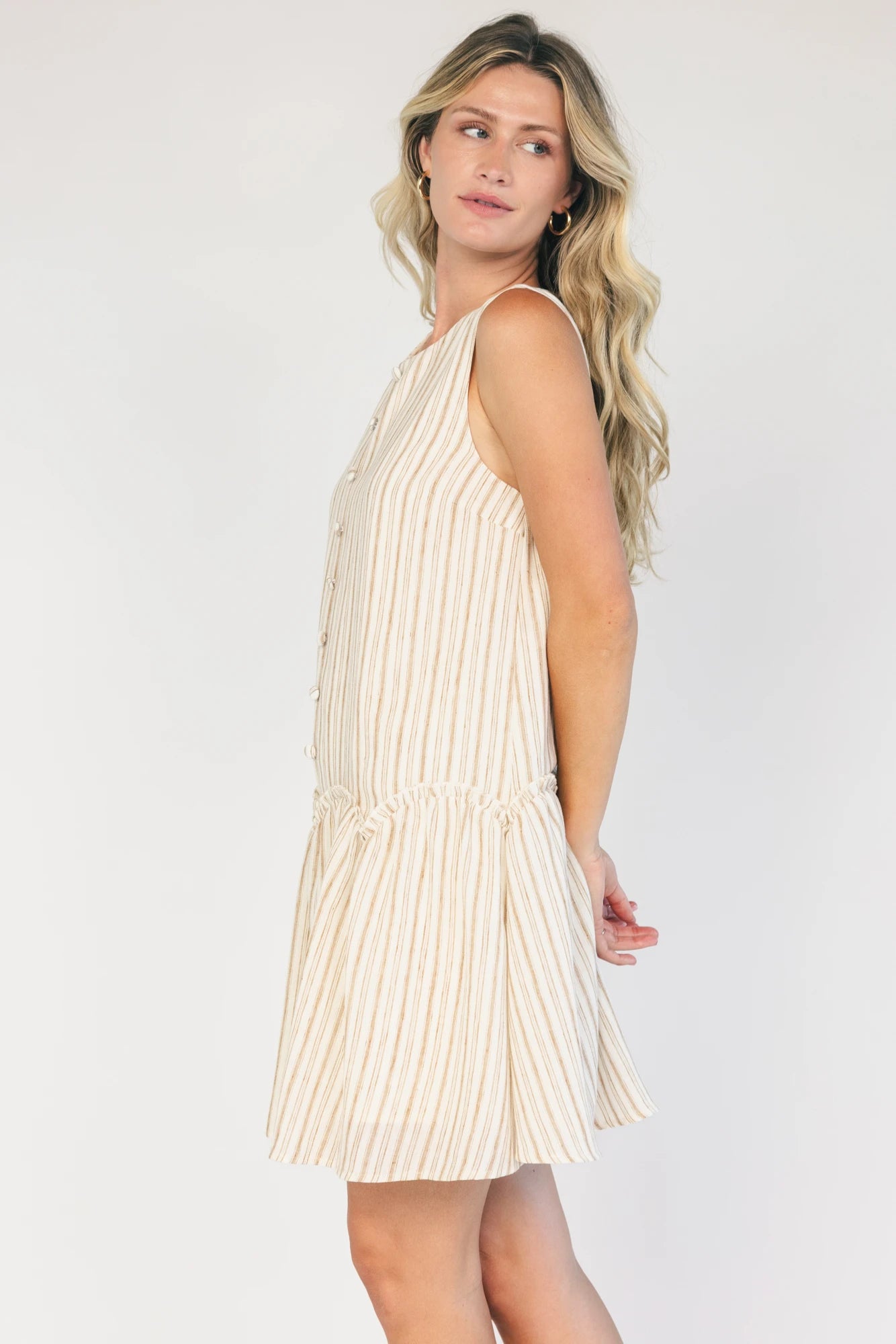 Model wearing an ivory and taupe striped sleeveless mini dress with a round neckline, faux button-front detail, and a curved drop-waist seam that flows into a gathered ruffle hem.