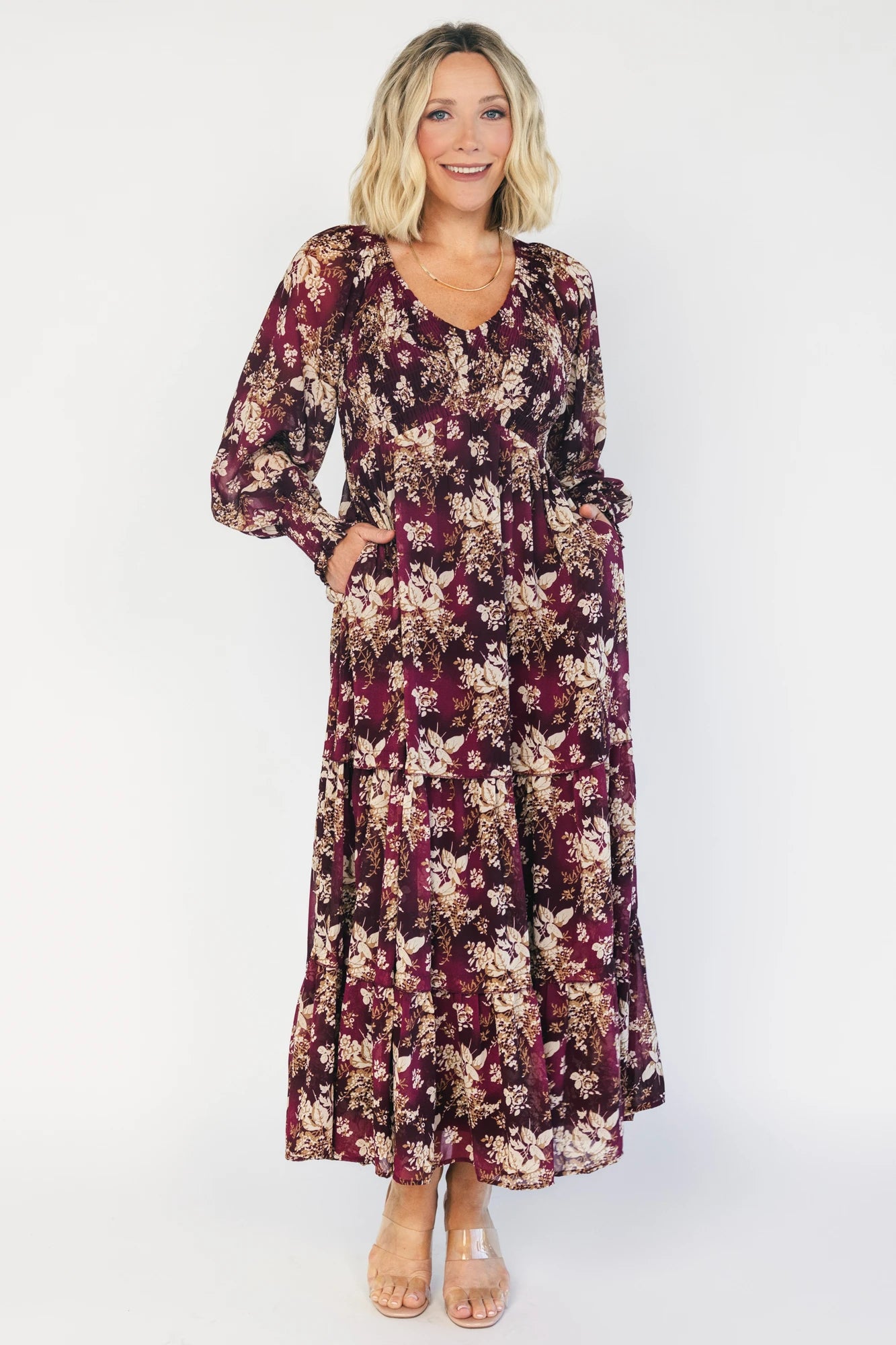 Model wearing a burgundy floral maxi dress with a V-neckline, smocked bodice, and long sheer sleeves with smocked ruffle cuffs.