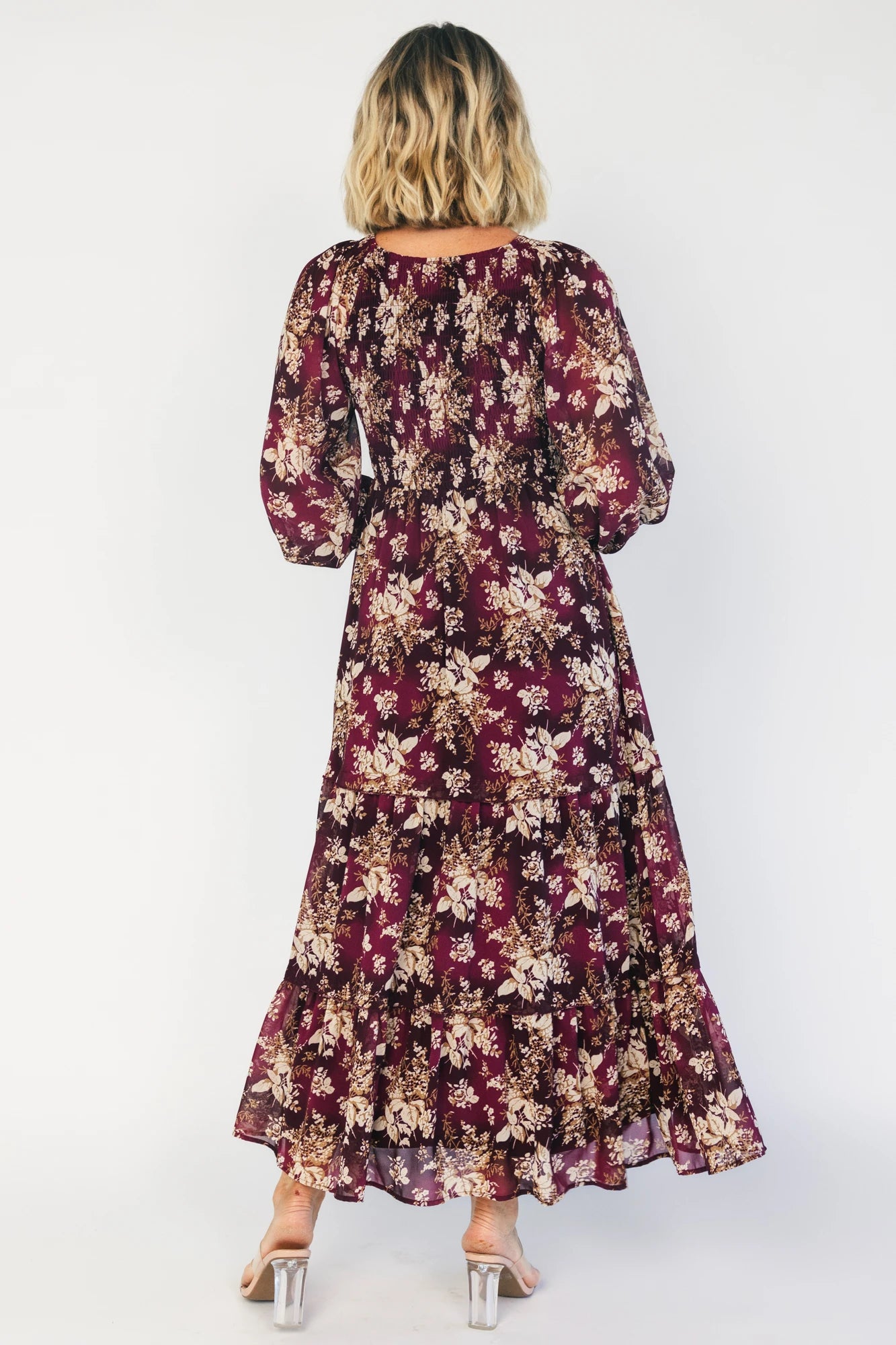 Model wearing a burgundy floral maxi dress with a V-neckline, smocked bodice, and long sheer sleeves with smocked ruffle cuffs.