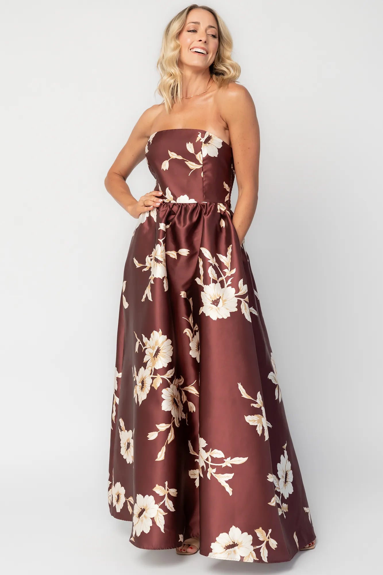 Model wearing a strapless maxi gown with a mahogany floral print and a fitted bodice. A rosette detail accents the neckline, while the flowing floor-length skirt creates elegant movement.