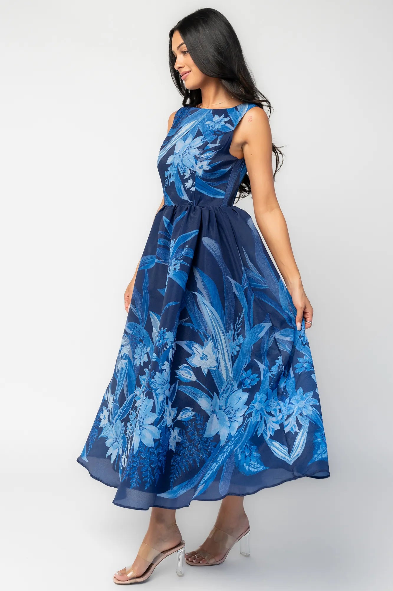 Model wearing a navy sleeveless midi dress with a high boat neckline and a fitted bodice. A tonal blue floral print adds subtle dimension throughout, while the defined waist flows into a full midi skirt that creates graceful movement and an elegant, timeless silhouette.