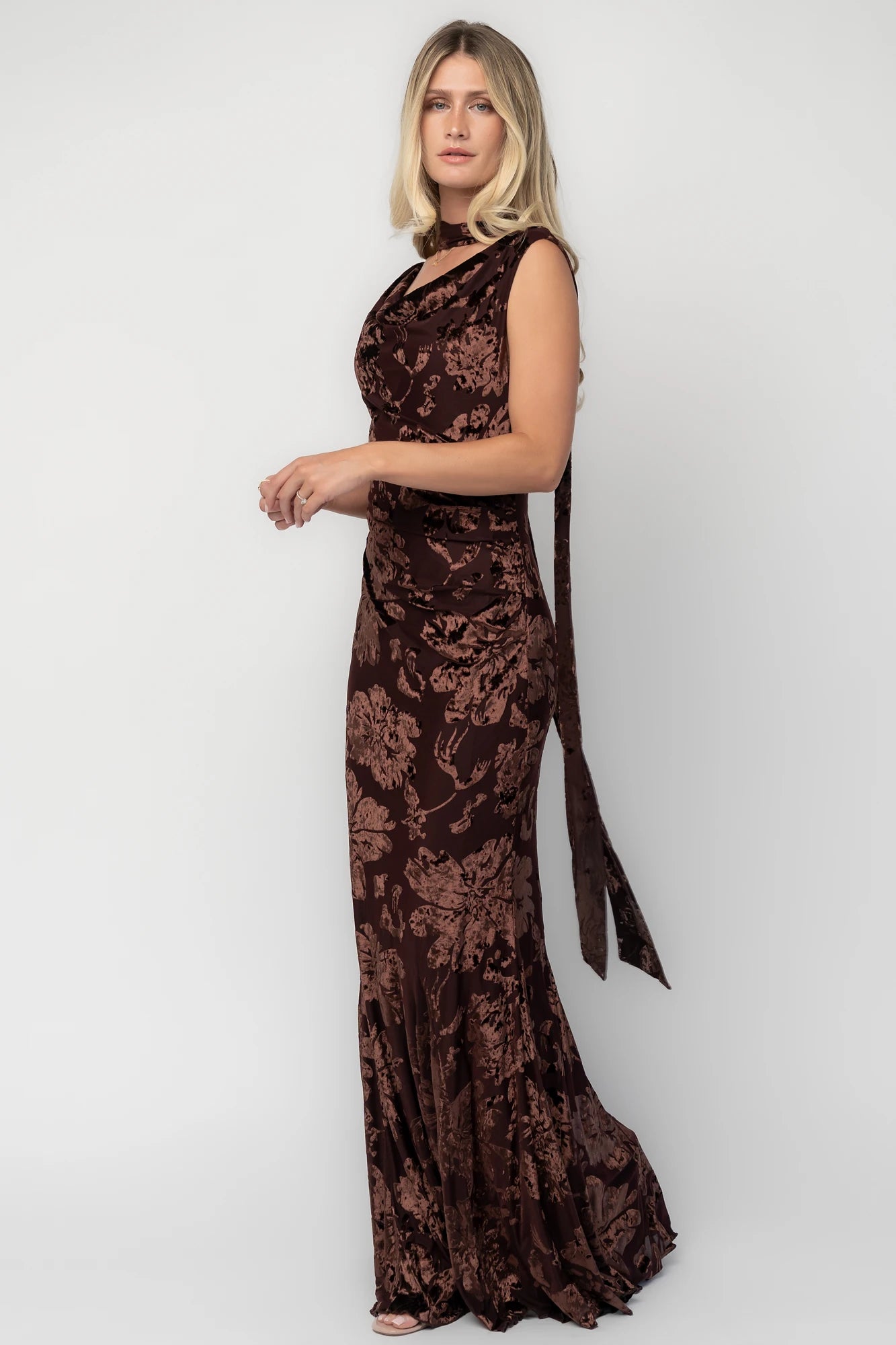 Model wearing a brown velvet burnout floral maxi dress with a draped cowl neckline, scarf detail, side ruching, and a flowing mermaid silhouette.