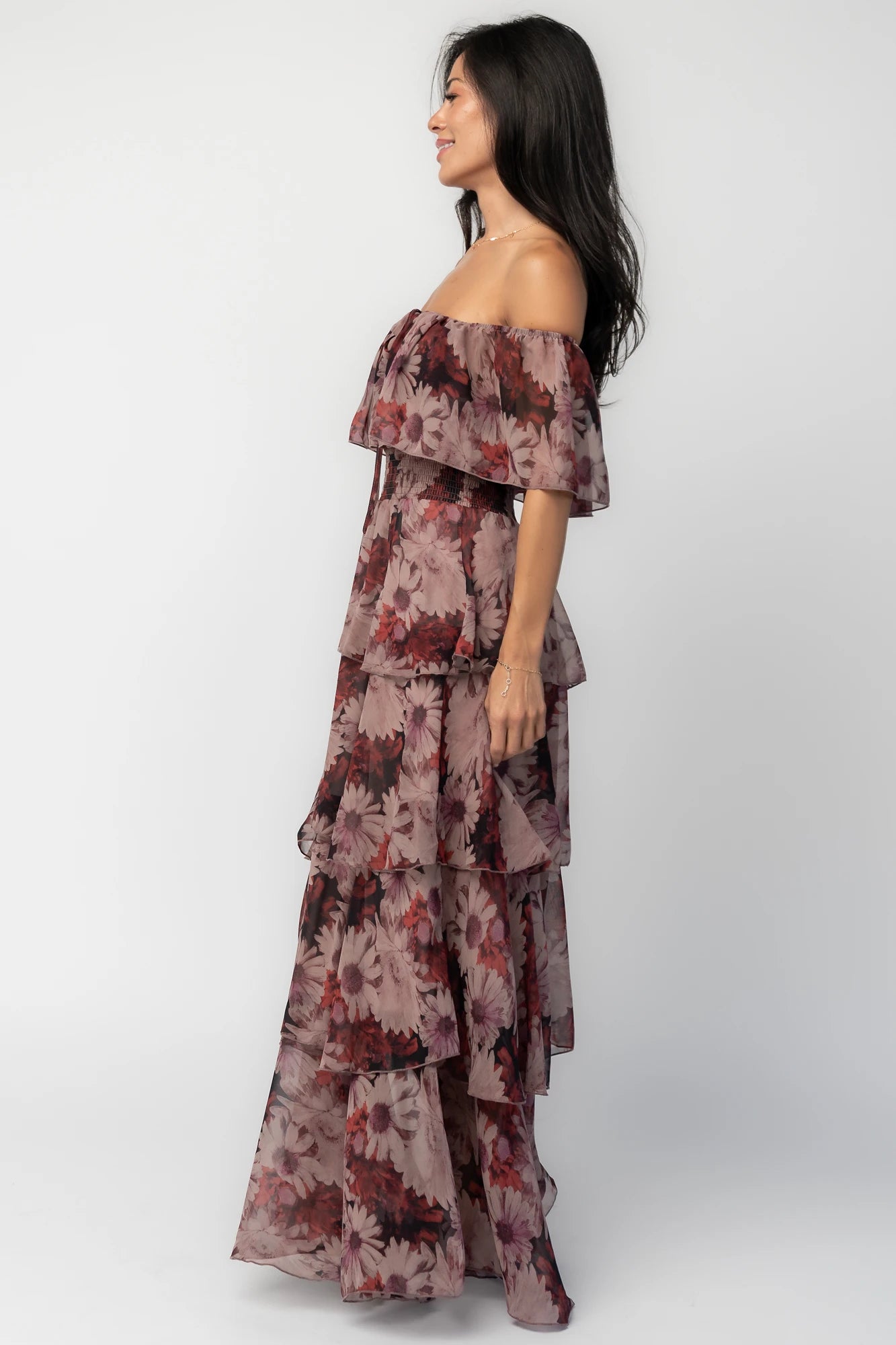 Model wearing the Carina Off-Shoulder Maxi in a soft multi floral print featuring a versatile neckline that can be worn on or off the shoulders, a ruffle overlay across the bust, a smocked waist, and a flowing four-tier maxi skirt.
