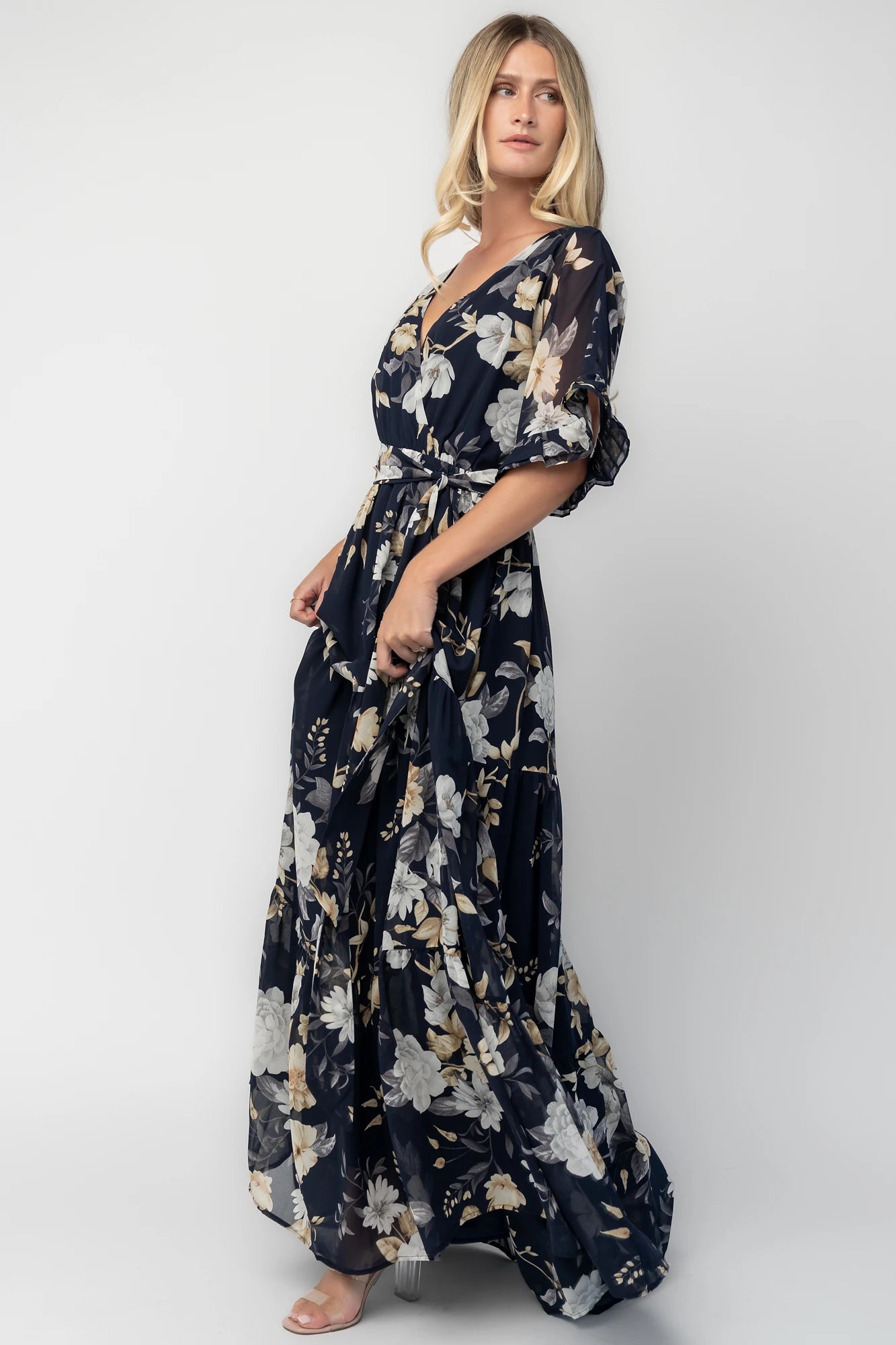 Model wearing the Luna Maxi Dress in Navy Floral featuring a surplice V-neckline with a V-back and tie detail, soft flutter sleeves, and an elastic waist with a removable sash tie