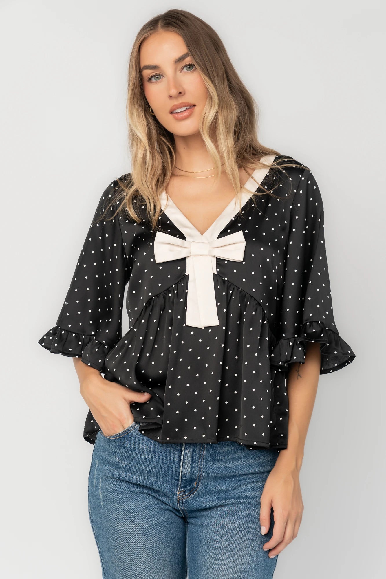 Black satin polka dot peplum top with an ivory contrast collar, decorative bow, V-neckline, and elbow-length ruffle sleeves.