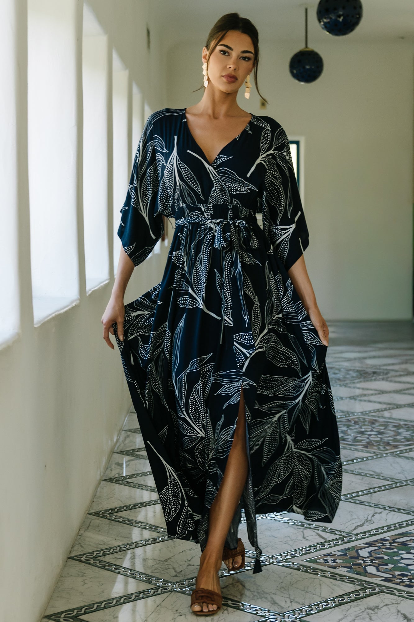 Model wearing Kalea navy maxi dress with off-white botanical print, surplice neckline and detached sash, full-length front view on studio background.