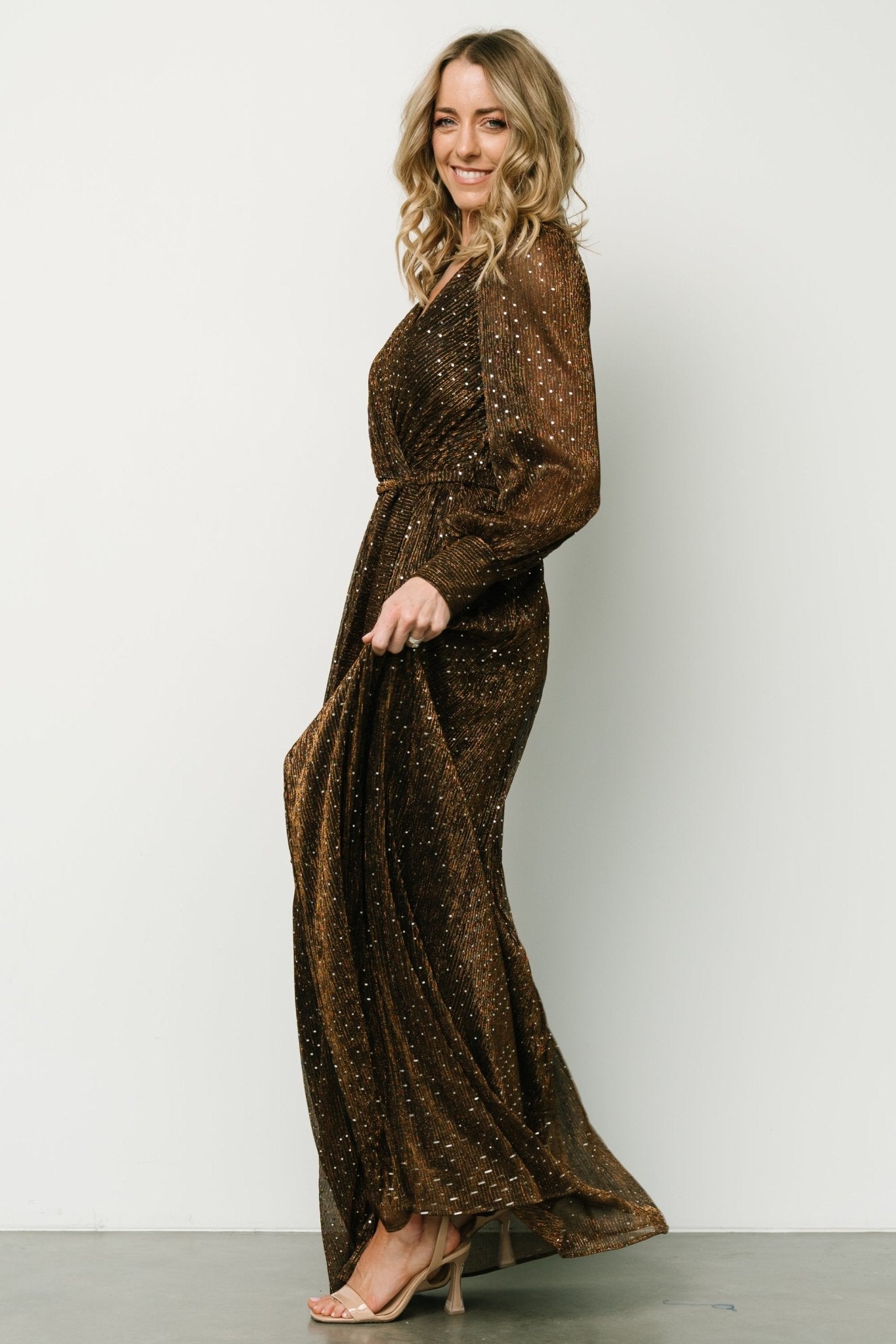 Side view highlighting the left-side leg slit and graceful drape of the bronze sequin maxi dress on a standing model.
