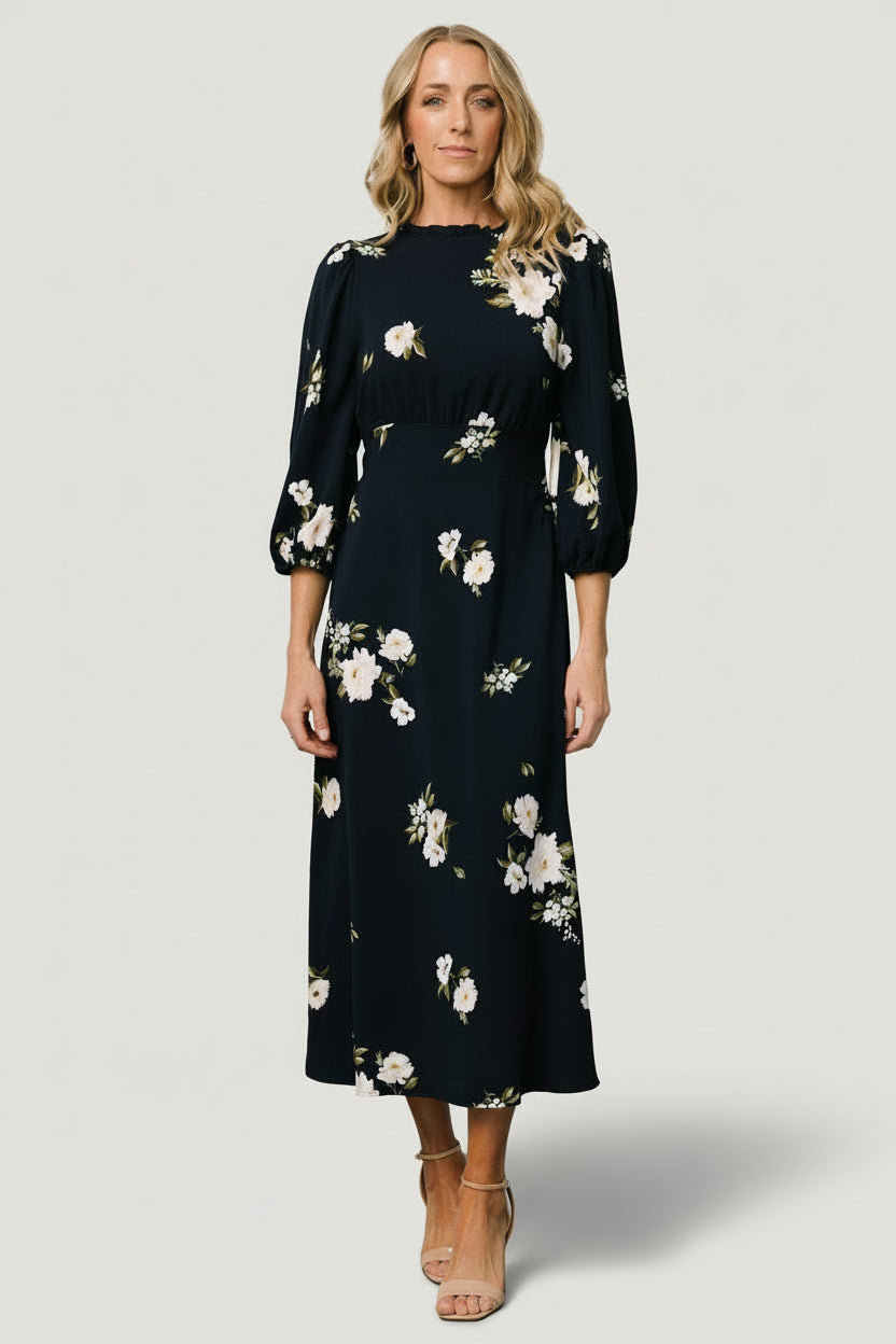 Styled shot of Karalie maxi dress on model, navy floral print, smocked waist, ruffled mock neck and mid-length sleeves visible.