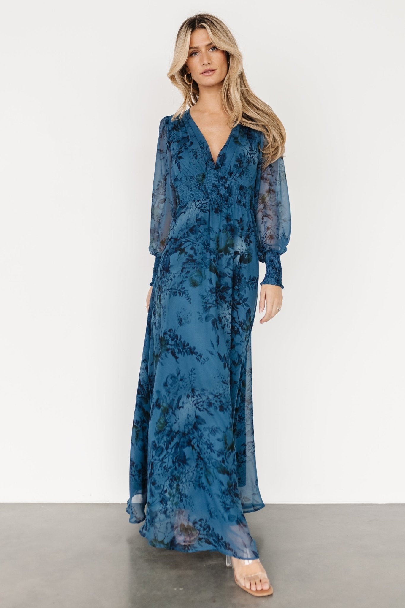 Model wearing deep blue and black floral chiffon maxi dress with deep V-neck, smocked waist and long smocked sleeves, front view.