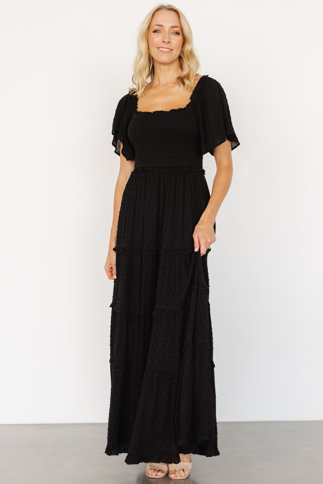 Three-quarter side view of black Lenora maxi dress on model; short flutter sleeves and ruffled elastic waist visible with soft drape of rayon fabric.