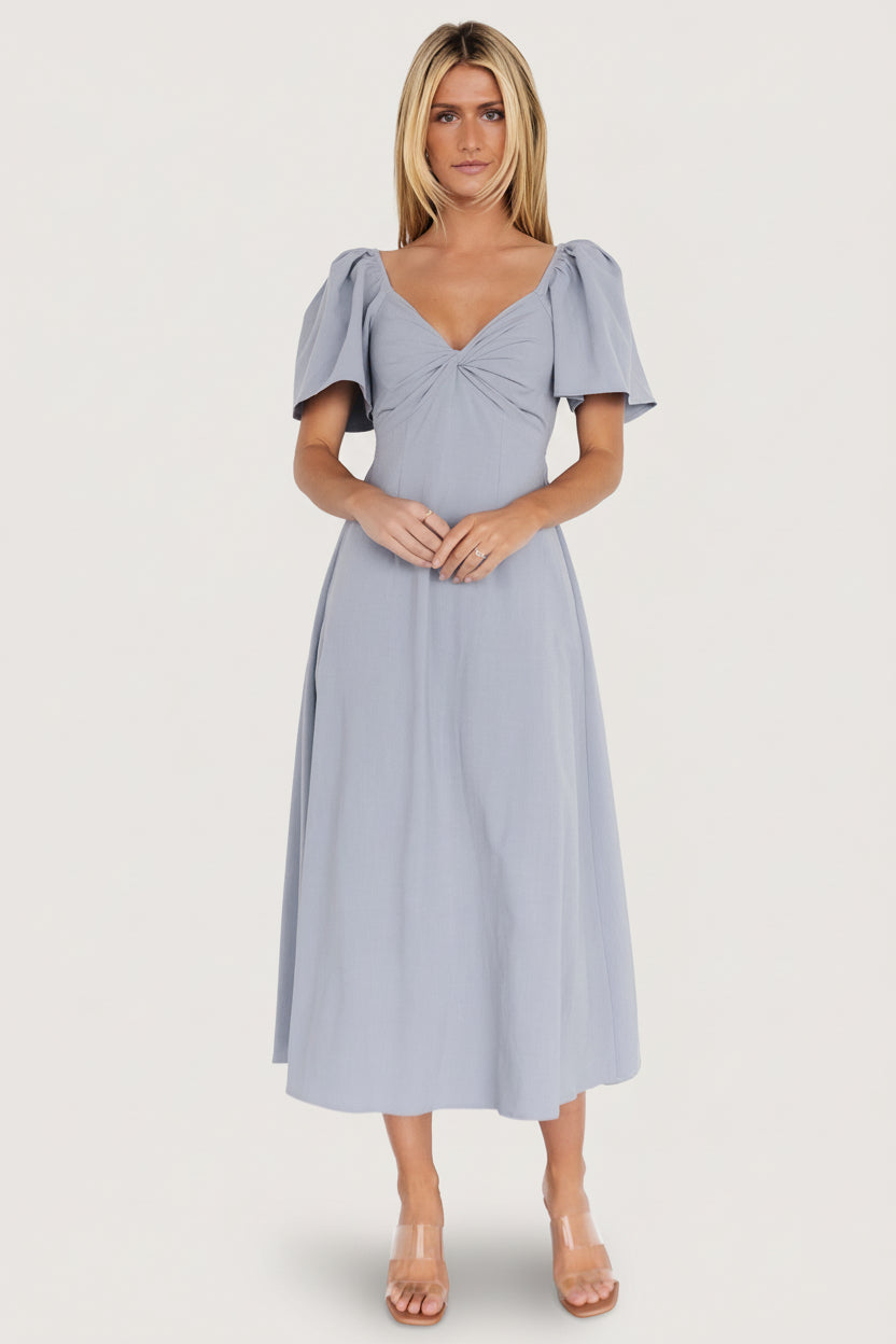 Model wearing dusty blue Indie Back Tie Dress walking forward; visible open back with adjustable tie and mid-length hem.