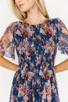 Model wearing the Mara dark blue multi floral tulle dress showing the smocked bodice, flutter sleeves, and tiered midi skirt in natural light.