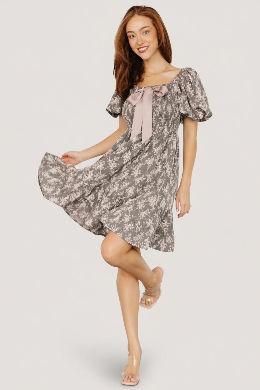 Model styling shot of the Anastasia Bow Short Dress in Dusty Olive Print, illustrating overall silhouette, short length, and soft polyester drape.