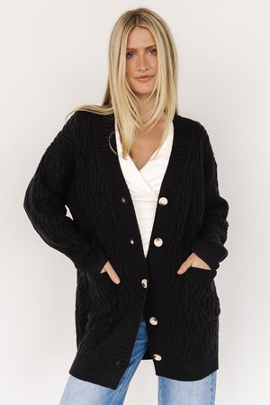 Front view of Alberta cable knit cardigan in black on model, showing chunky cable texture, dropped shoulders, wide ribbed placket, and five-button closure.