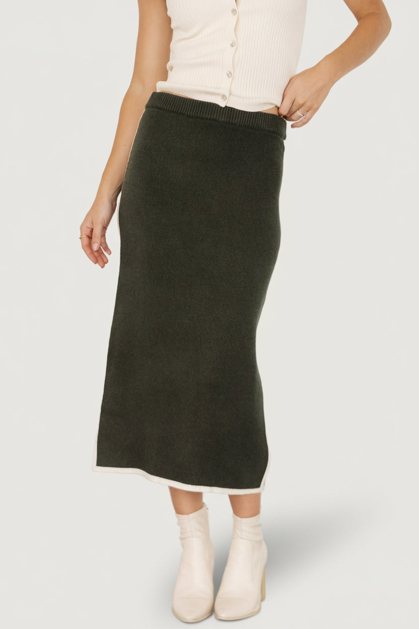 Front view of Ava Knit Midi Skirt in dark green on model, showing midi length, elastic waistband, and cream piping detail at waist and hem.