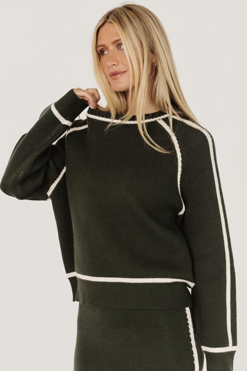 Front view of model wearing dark green Ava Knit Sweater, showing round neckline, cream piping, and relaxed tailored silhouette.