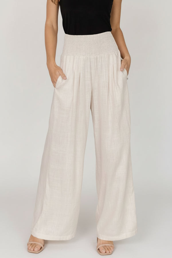 Front view of model wearing Parvati smocked high-waist wide-leg pants in natural linen-rayon blend, showing smocked waist and side pockets.
