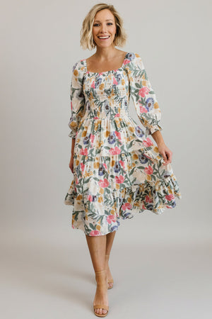Front view of a smocked midi dress in muted multi floral print on a model, showing square neckline and tiered skirt, full-length.