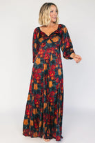 Model wearing a dark jade maxi dress with an oversized marsala floral print, a twist-knot V-neckline, half sleeves with elastic cuffs, a defined high waist, and a flowing maxi skirt with godet panels and an asymmetrical scalloped hem.