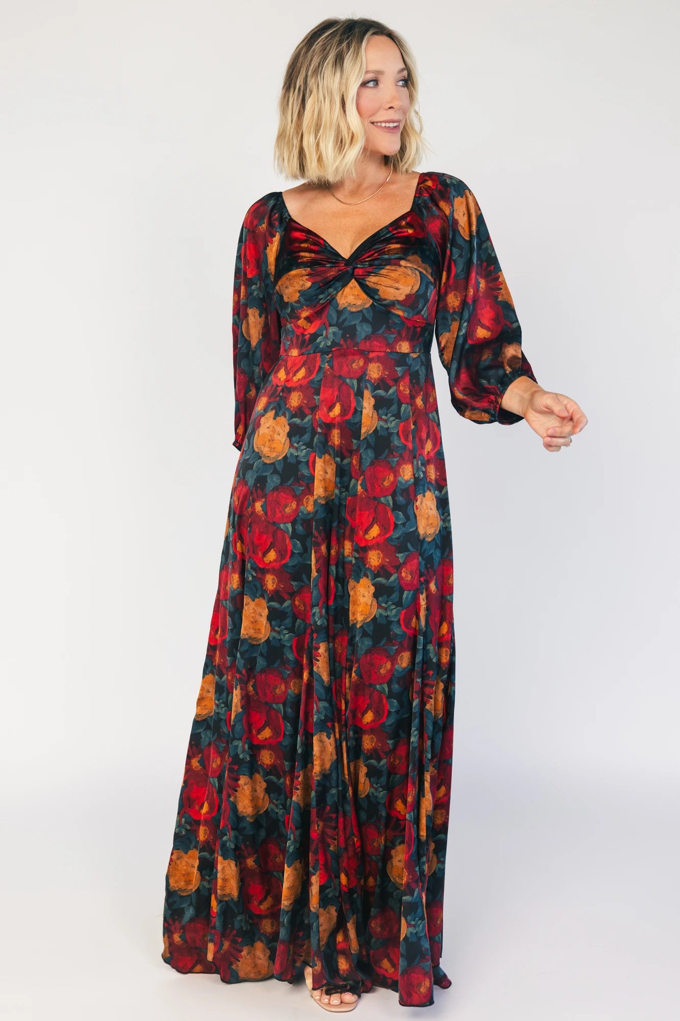 Model wearing a dark jade maxi dress with an oversized marsala floral print, a twist-knot V-neckline, half sleeves with elastic cuffs, a defined high waist, and a flowing maxi skirt with godet panels and an asymmetrical scalloped hem.