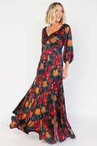 Model wearing a dark jade maxi dress with an oversized marsala floral print, a twist-knot V-neckline, half sleeves with elastic cuffs, a defined high waist, and a flowing maxi skirt with godet panels and an asymmetrical scalloped hem.