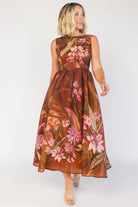 Model wearing a brown floral midi dress with a high boat neckline, sleeveless fitted bodice, and defined waist. The dress features a pink floral print throughout and a flowing midi-length skirt, creating an elegant, timeless silhouette.