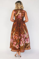 Model wearing a brown floral midi dress with a high boat neckline, sleeveless fitted bodice, and defined waist. The dress features a pink floral print throughout and a flowing midi-length skirt, creating an elegant, timeless silhouette.