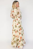 Model wearing a cream floral maxi dress with a surplice V-neckline, long sheer sleeves with elastic cuffs, and an elastic back waist with attached side ties. The chiffon dress features a cream base with rust and green florals, an angled tiered skirt, and a flowing silhouette with a midi-length lining.