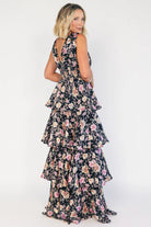 Model wearing a black floral maxi dress with a deep V-neckline, banded waist, and cascading tiered skirt. The dress features a black background with pink, yellow, and orange floral print, creating a flowing, romantic silhouette.