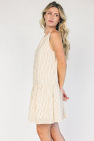 Model wearing an ivory and taupe striped sleeveless mini dress with a round neckline, faux button-front detail, and a curved drop-waist seam that flows into a gathered ruffle hem.