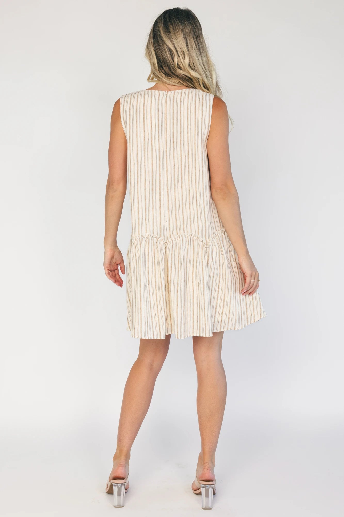 Model wearing an ivory and taupe striped sleeveless mini dress with a round neckline, faux button-front detail, and a curved drop-waist seam that flows into a gathered ruffle hem.