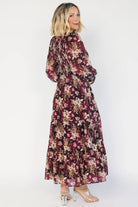 Model wearing a burgundy floral maxi dress with a V-neckline, smocked bodice, and long sheer sleeves with smocked ruffle cuffs.