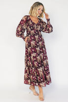 Model wearing a burgundy floral maxi dress with a V-neckline, smocked bodice, and long sheer sleeves with smocked ruffle cuffs.