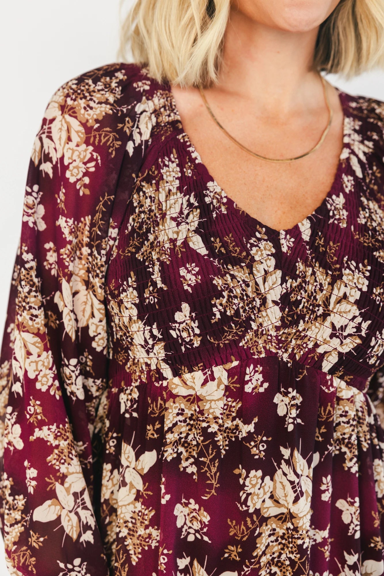 Model wearing a burgundy floral maxi dress with a V-neckline, smocked bodice, and long sheer sleeves with smocked ruffle cuffs.