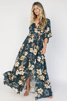 Model wearing a dark jade floral maxi dress with a surplice V-neckline, kimono sleeves, and an elastic waist with a removable sash tie.