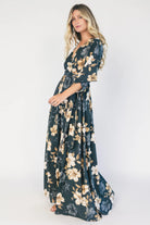 Model wearing a dark jade floral maxi dress with a surplice V-neckline, kimono sleeves, and an elastic waist with a removable sash tie.