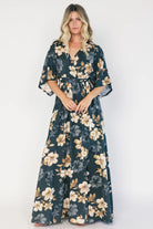 Model wearing a dark jade floral maxi dress with a surplice V-neckline, kimono sleeves, and an elastic waist with a removable sash tie.