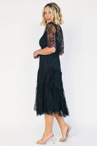 Model wearing a black lace midi dress with a deep V-neckline, short sheer scalloped sleeves, and an elastic waist.