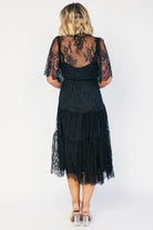 Model wearing a black lace midi dress with a deep V-neckline, short sheer scalloped sleeves, and an elastic waist.