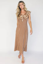 Model wearing a dusty mocha and cream plaid midi dress with a V-neckline, decorative bow tie detail, cascading ruffle trim, and a fitted silhouette.