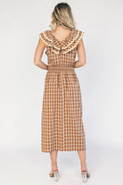 Model wearing a dusty mocha and cream plaid midi dress with a V-neckline, decorative bow tie detail, cascading ruffle trim, and a fitted silhouette.