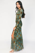 Model wearing a green mesh maxi dress with copper velvet burnout detailing, a high round neckline, and long sheer sleeves.