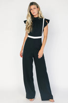 Model wearing the Lumina Ruffle Jumpsuit in Black + Ivory featuring a sleeveless ruffle-trim bodice, defined waist, and flowing wide-leg pants.