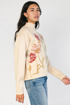 Natural knit sweater with a mock neckline, long sleeves, relaxed fit, and an embroidered floral motif at the neckline.