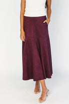 Model wearing a mulberry faux suede A-line midi skirt with a high-rise waistband, stitched panel detailing, side pockets, and a raw hem for a timeless, feminine silhouette.