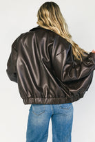 Model wearing a dark brown faux leather bomber jacket with a folded lapel collar, front zipper closure, elastic hem, silver snap cuffs, and front pockets.