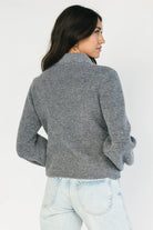 Model wearing a gray knit cardigan with a scalloped collar, adjustable black velvet bow detail, two front hook-and-eye closures, and a relaxed fit.
