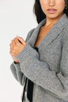 Model wearing a gray knit cardigan with a scalloped collar, adjustable black velvet bow detail, two front hook-and-eye closures, and a relaxed fit.