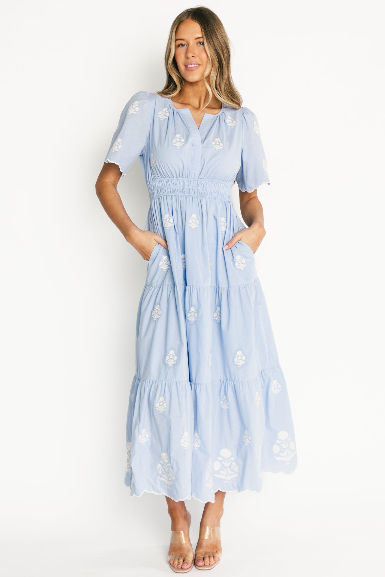 Model wearing the Delilah Embroidered Maxi Dress in light blue, front view showing split V-neck, scalloped short sleeves, embroidered off-white floral pattern and smocked waist.